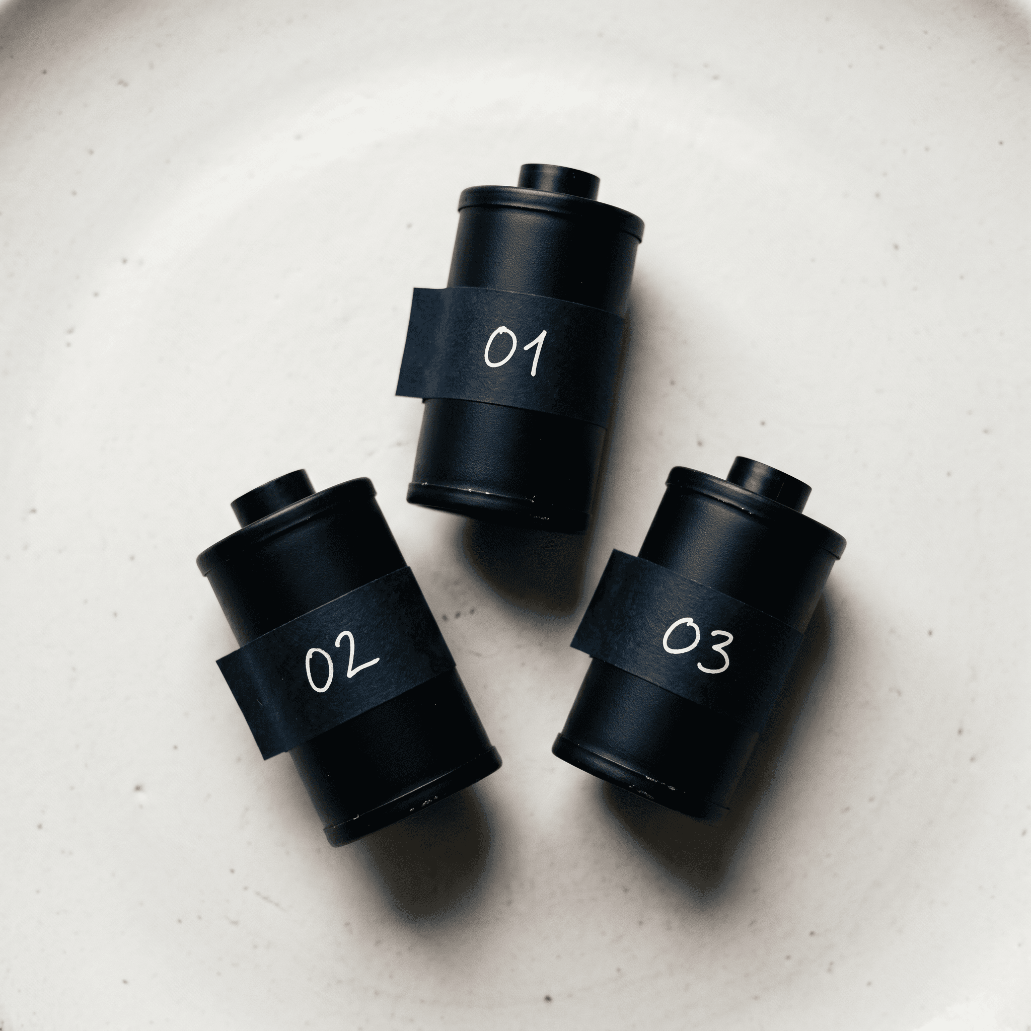 Editorial overhead flat lay of three matte film canisters in a triangular arrangement on a warm white Nordic surface representing Kling, Pika, and Luma video models